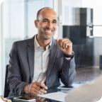 Man wearing a gray suit smiles and sits at a glass-top desk with a pen and paper.